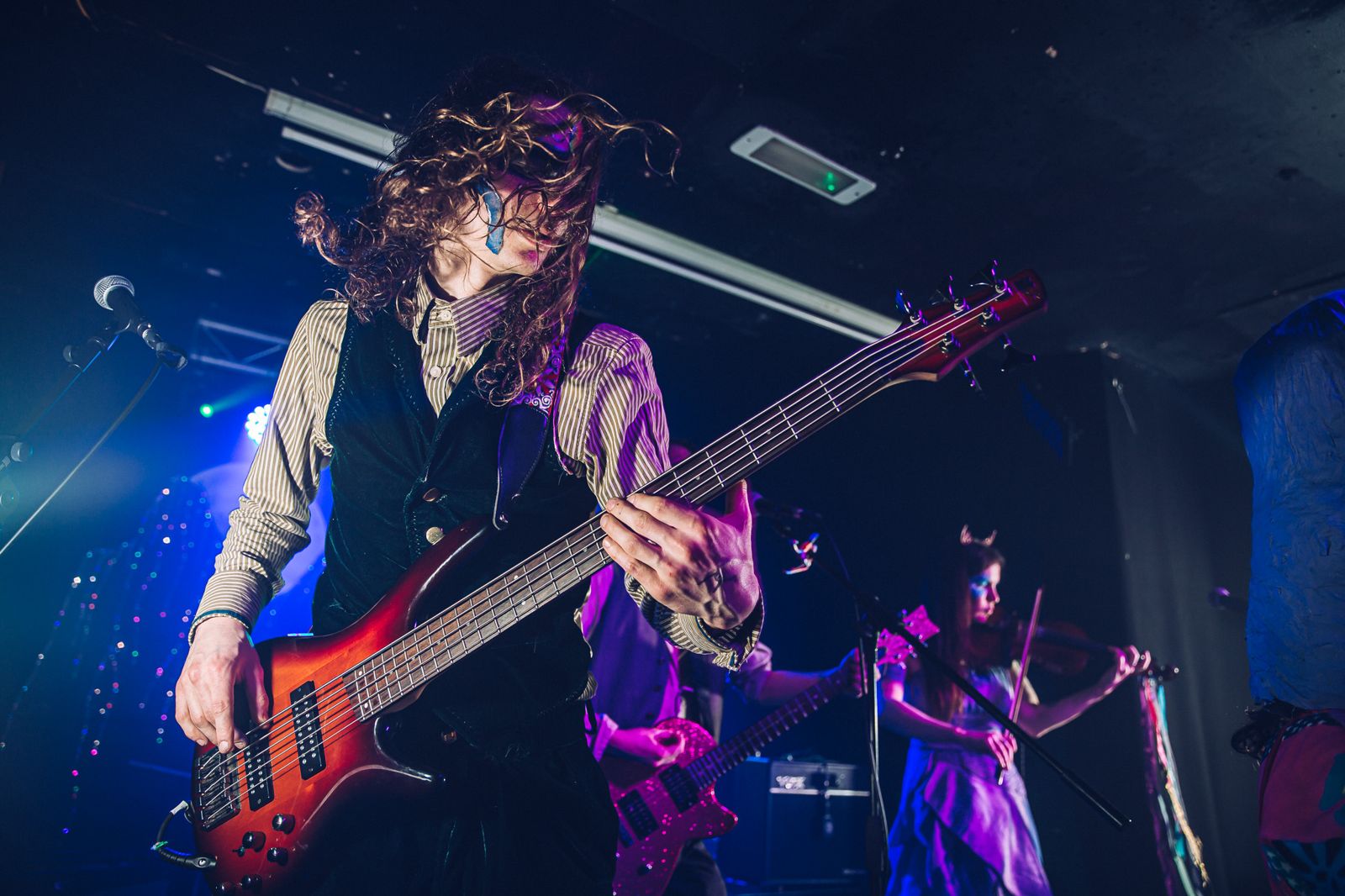 Bassist with long curly hair playing a red five-string bass under blue stage lights, with a violinist behind.