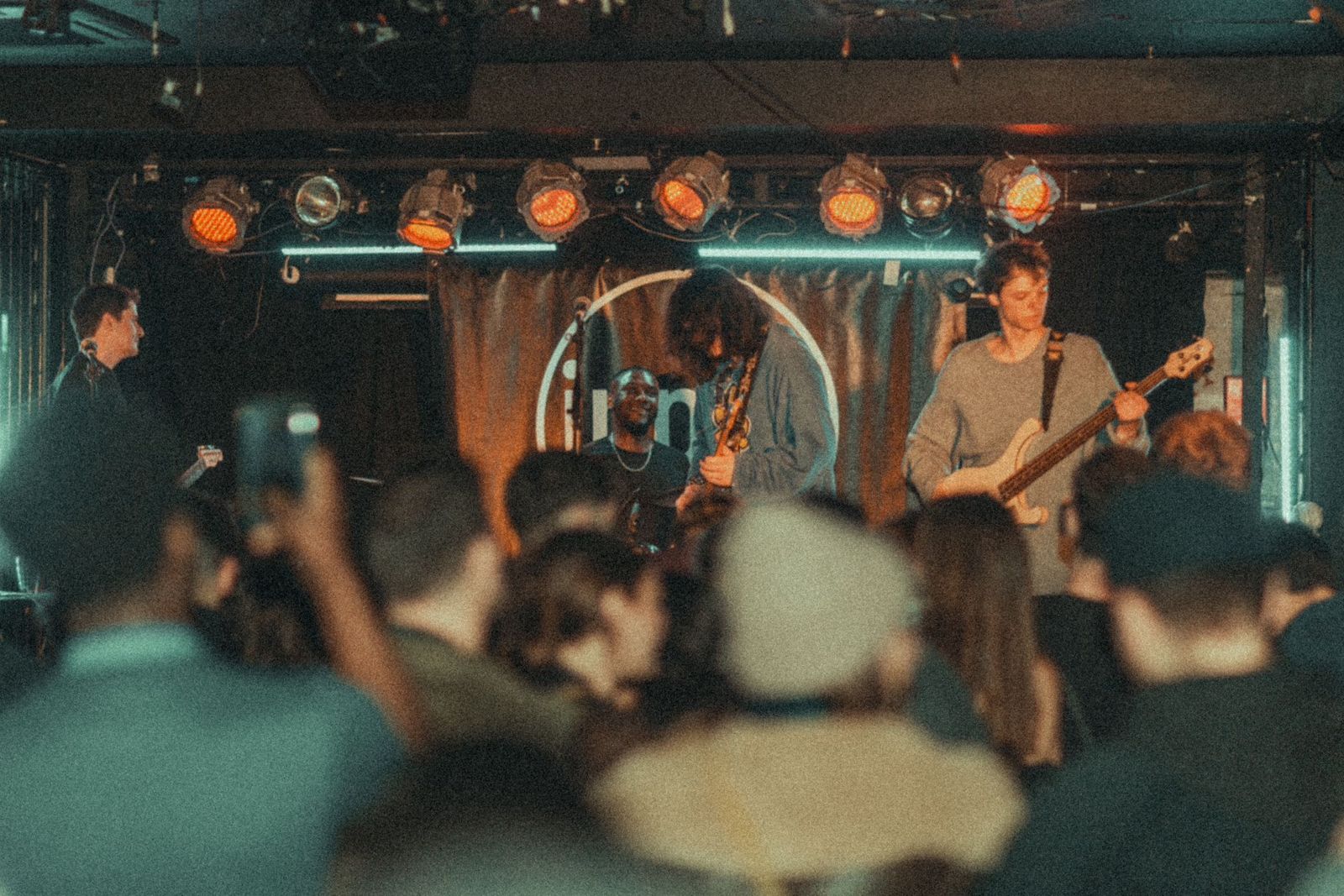 A band playing on a small venue stage under a row of orange lights, seen over the heads of the crowd.